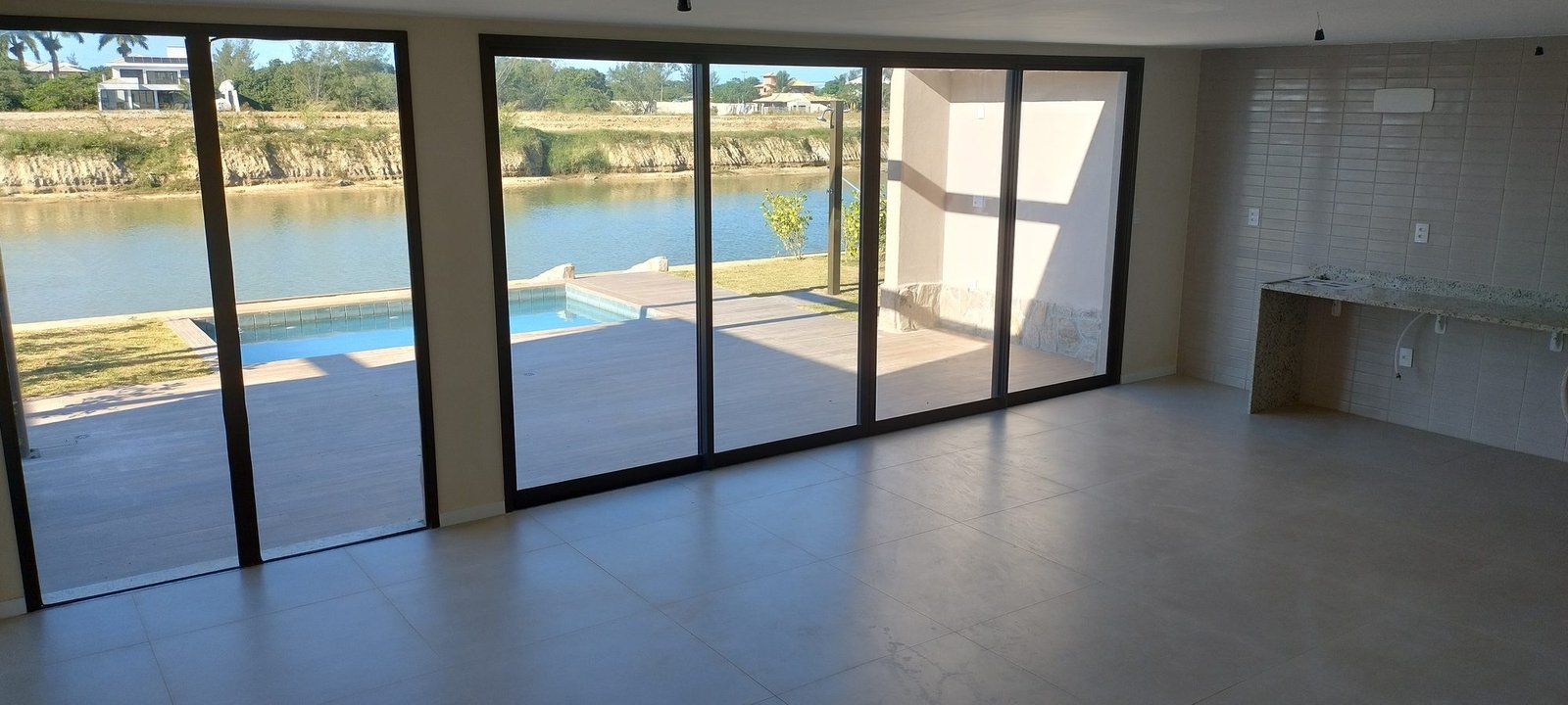 phoco-sync-ec2dfcf94ac593c0-b2427f50cf9a Ambiente sem móveis de uma casa do Toriba Sul, com portas de vidro abertas para a piscina e o canal (foto real)