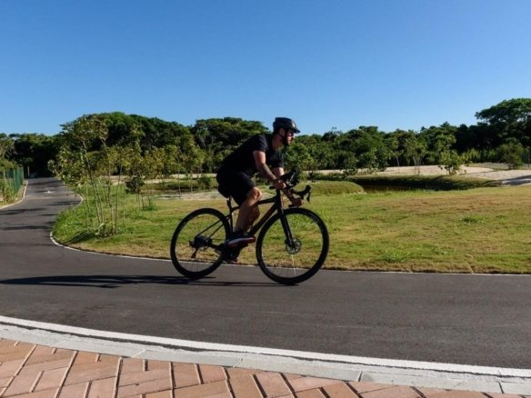 Ciclista pedalando numa via asfaltada do Aretê Búzios, com gramado e árvores ao lado (foto real)