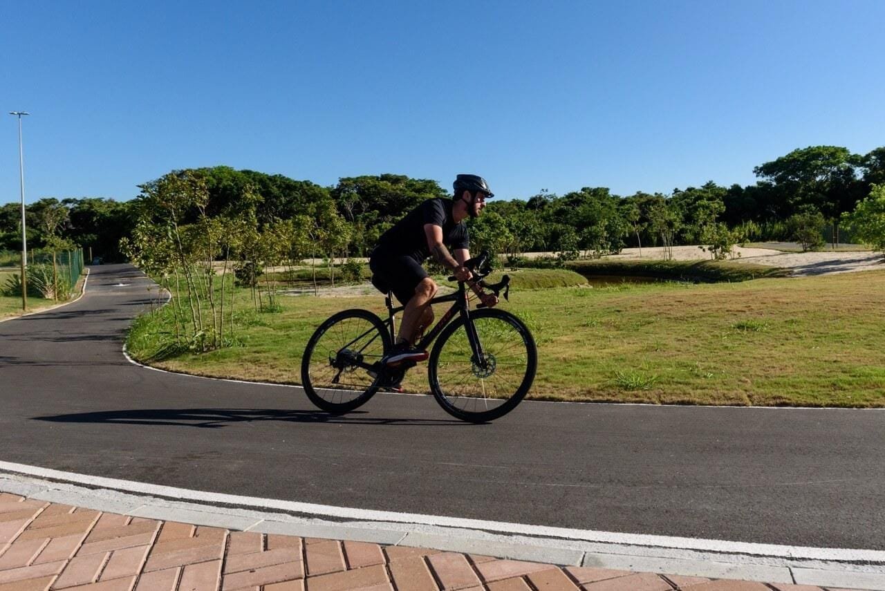 Ciclista pedalando numa via asfaltada do Aretê Búzios, com gramado e árvores ao lado (foto real)