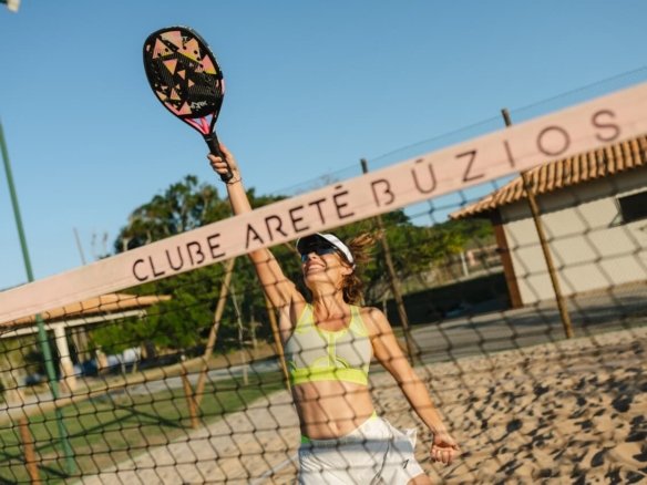 Jogadora de beach tennis na quadra de areia, com a placa do Clube Aretê Búzios ao fundo (foto real)