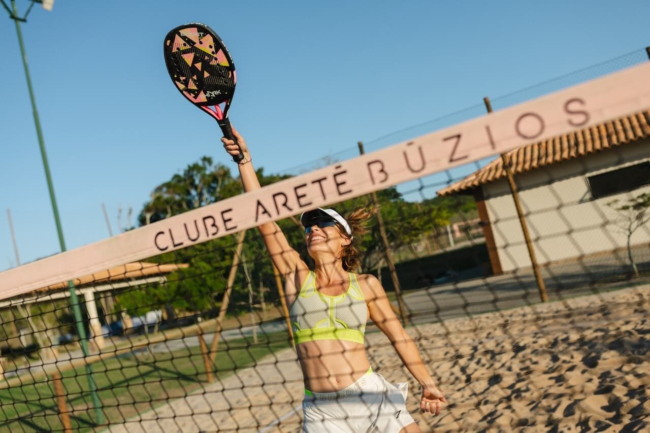 Jogadora de beach tennis na quadra de areia, com a placa do Clube Aretê Búzios ao fundo (foto real)