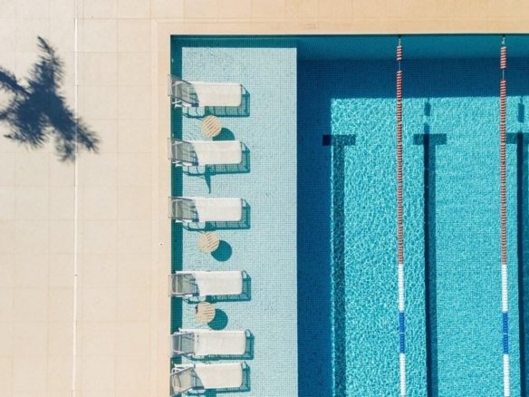 Piscina de raias vista de cima, com espreguiçadeiras na borda e a sombra de uma palmeira (foto real)