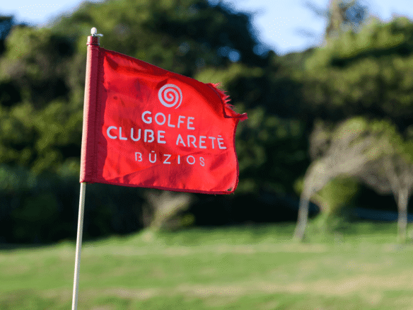 Bandeira vermelha do Golfe Clube Aretê Búzios no campo, com árvores ao fundo (foto real)