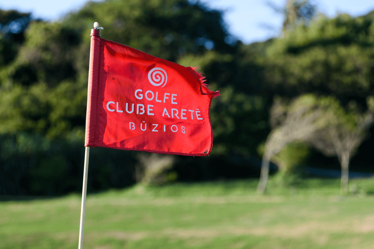 Bandeira vermelha do Golfe Clube Aretê Búzios no campo, com árvores ao fundo (foto real)