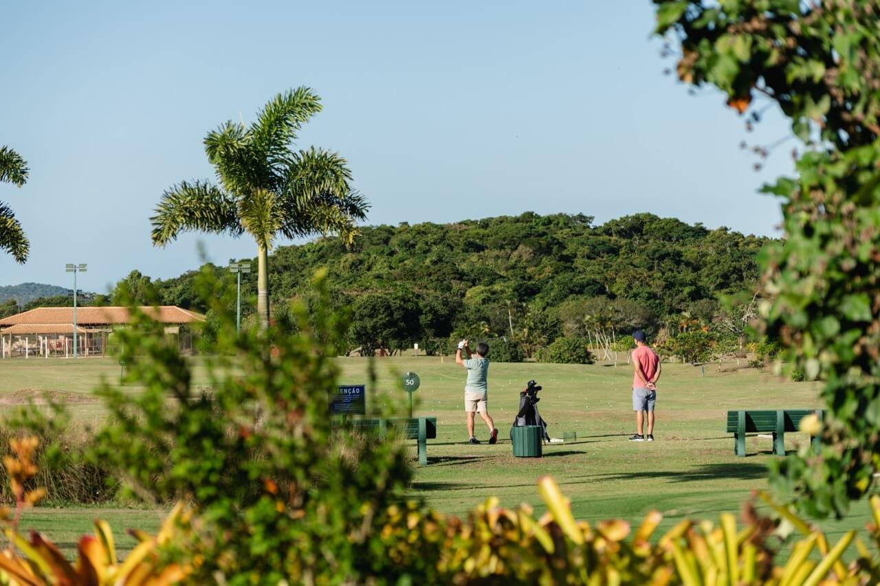 Dois jogadores de golfe no tee, com palmeiras e o morro ao fundo, vistos por entre plantas (foto real)