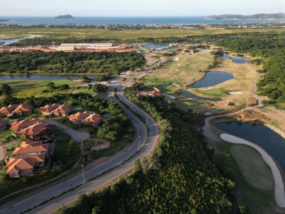 Vista aérea do Ybirá em maio de 2026, com casas à esquerda, a estrada e o campo de golfe com lagos, e o mar ao fundo (foto real)