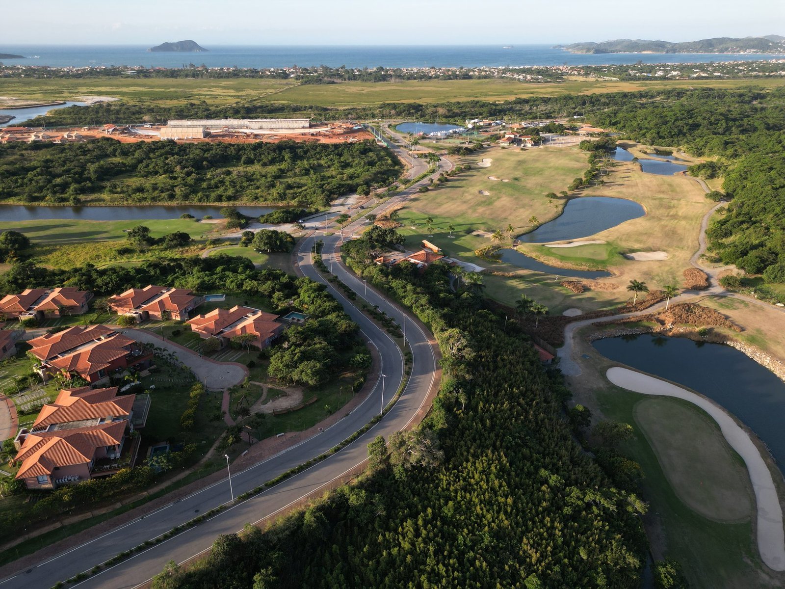 Vista aérea do Ybirá em maio de 2026, com casas à esquerda, a estrada e o campo de golfe com lagos, e o mar ao fundo (foto real)