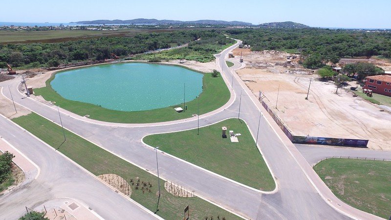 Vista aérea de um lago oval cercado de gramado, com avenidas e postes em volta, um terreno de terra em obras à direita e o mar e morros ao fundo.