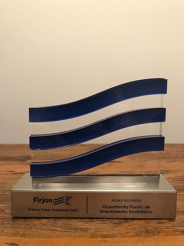 Troféu com três faixas azuis onduladas sobre uma base de metal, com as inscrições Firjan, Prêmio Firjan Ambiental 2021, Água e Efluentes e Opportunity Fundo de Investimento Imobiliário.