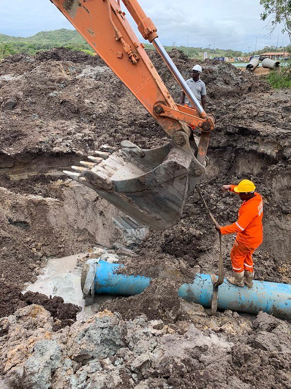 Vala aberta na terra com um tubo azul no fundo, de onde sai água. Um operário de macacão laranja e capacete amarelo está em pé sobre o tubo, segurando uma cinta presa à pá de uma escavadeira, e outro, de capacete branco, observa do alto do monte de terra.